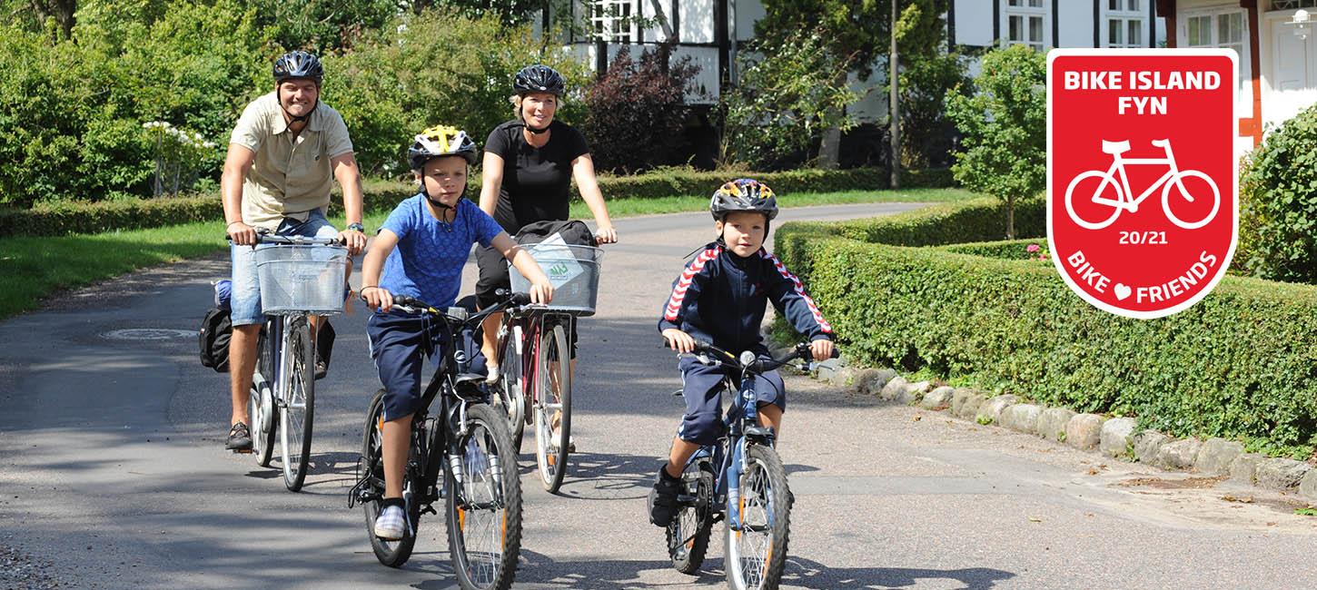 To voksne og to børn cykler af sted på stille asfaltvej med bindingsværkshus i baggrunden. Øverst i højre hjørne ses det røde Bike Friend-logo.