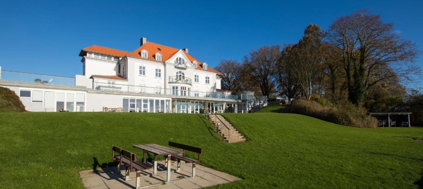 Stort, hvidt hotel med rødt tag beliggende i en stor have. Bænkesæt i forgrunden.