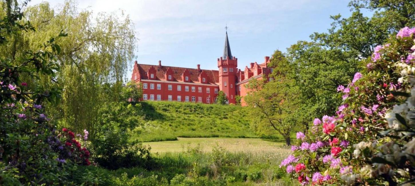 Rødt slot ses på afstand med flot spir og omringet af grønne buske og blomster