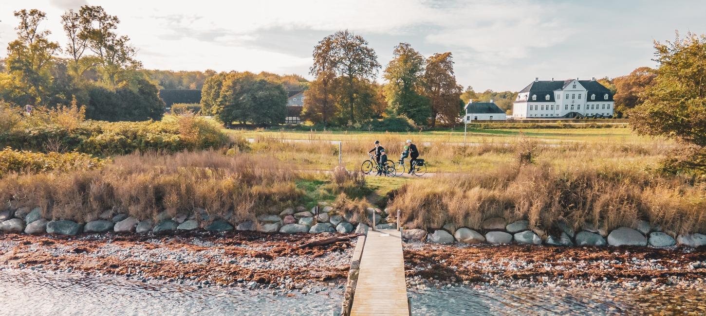 To cyklister gør holdet ved en badebro på strand med dige af store kampesten. I baggrunden en stor, hvid herregård og træer.