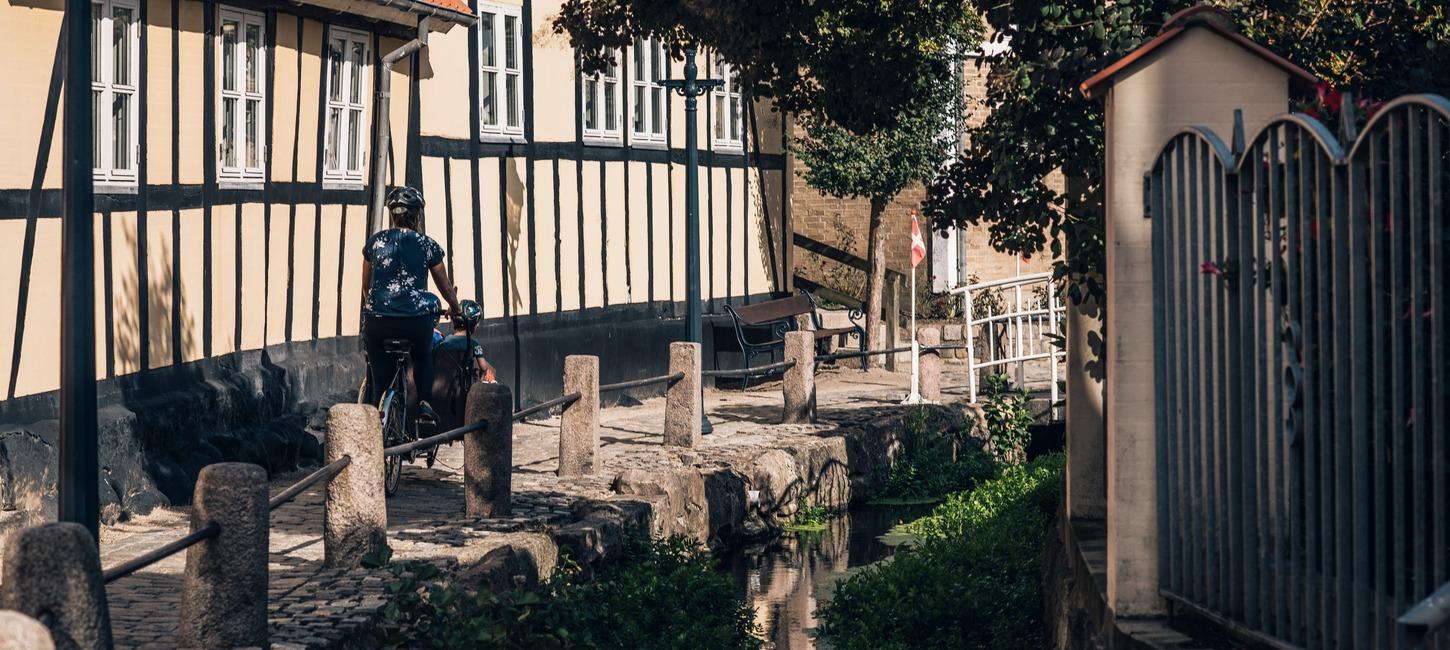 Cyklist trækker af sted med sin cykel på brostensbelagt sti langs med gult bindingsværkshus.