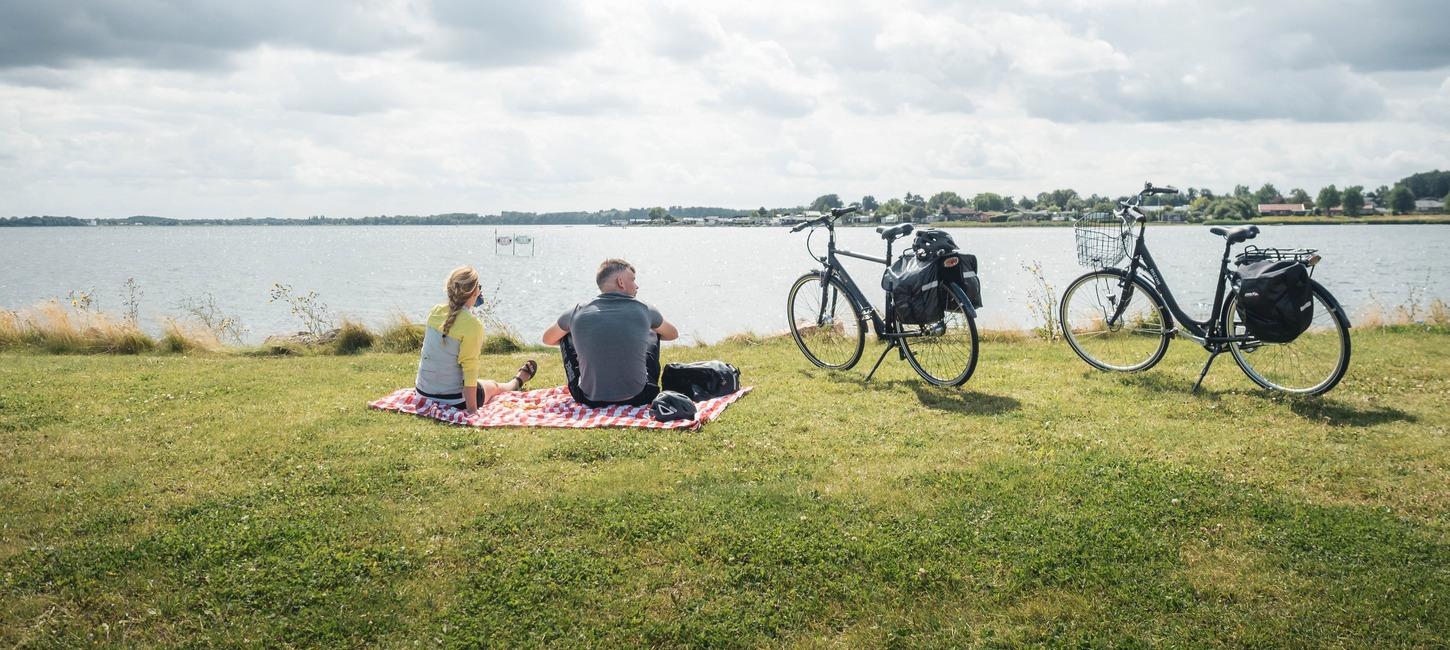 En mand og en kvinder holder rest på et ternet tæppe på græsset med udsigt over vandet. Deres cykler med oppakning står parkeret ved siden af.