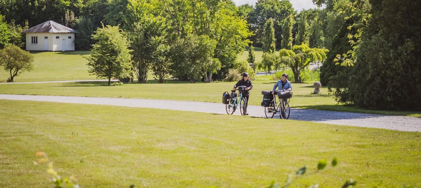 En par med hver deres cykel med opbakning går på en grussti. Foran stien er der græsplæne mens der bagved er græs og træer. 