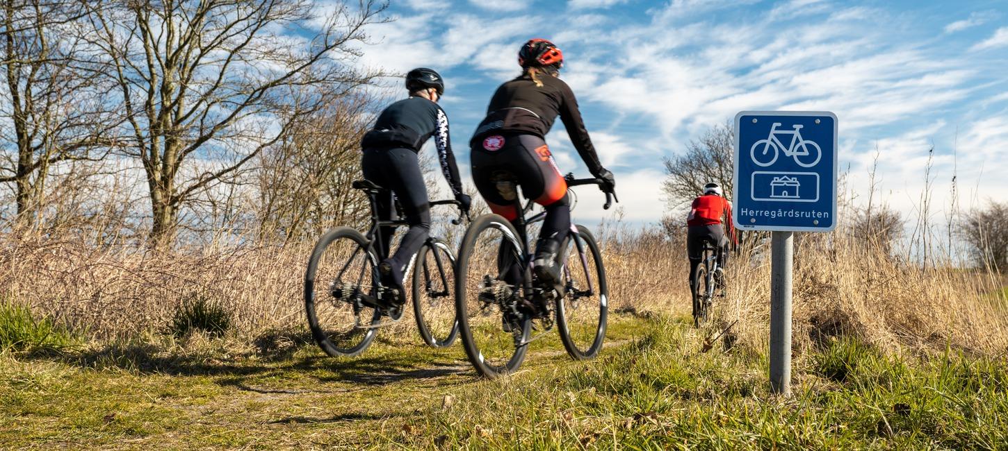 Tre racer-cyklister cykler på en græssti mellem højt græs og træer. I højre side ses et blåt skilt med cykel og navnet Herregårdsruten på.