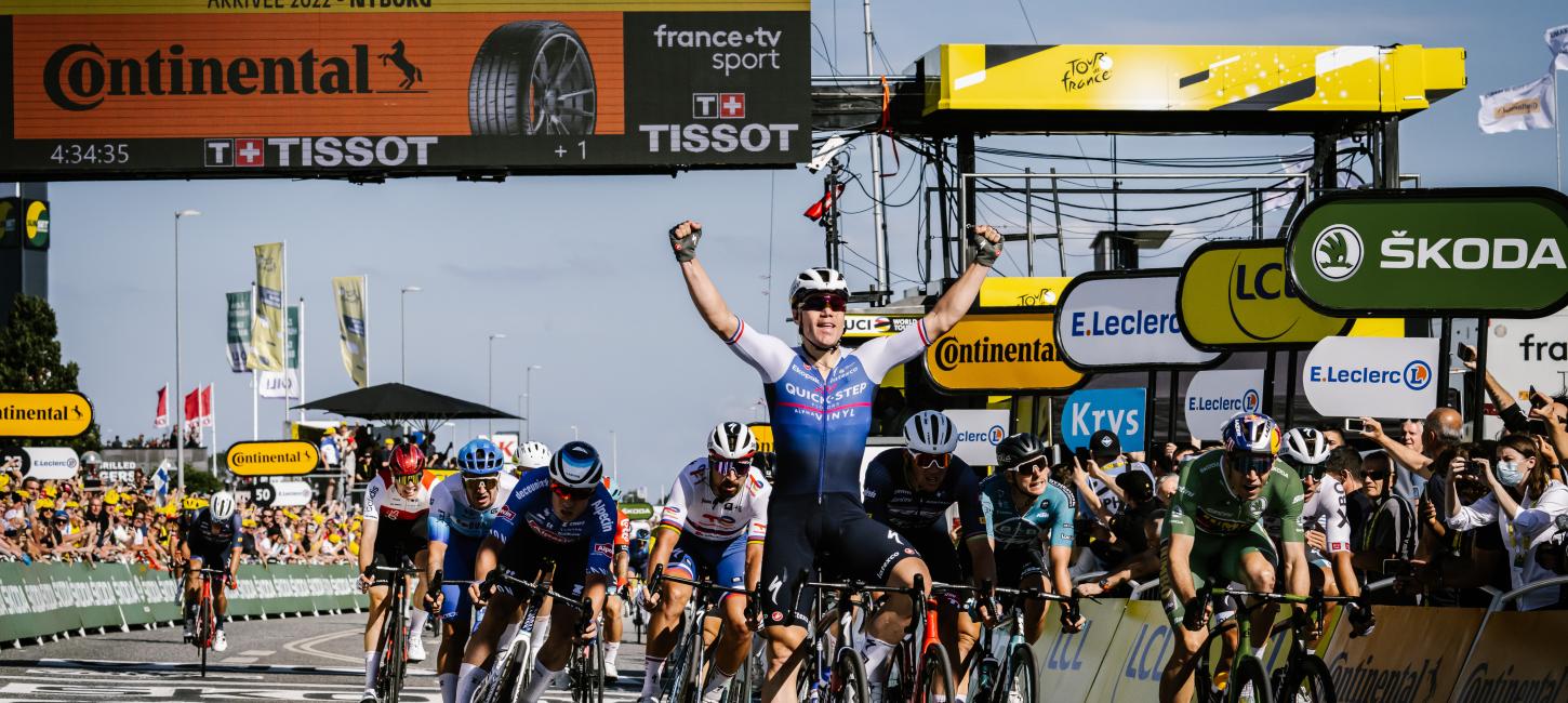 En masse cykelryttere til Tour de France fotograferet ved målområdet. En rytter har armene oppe i sejr.