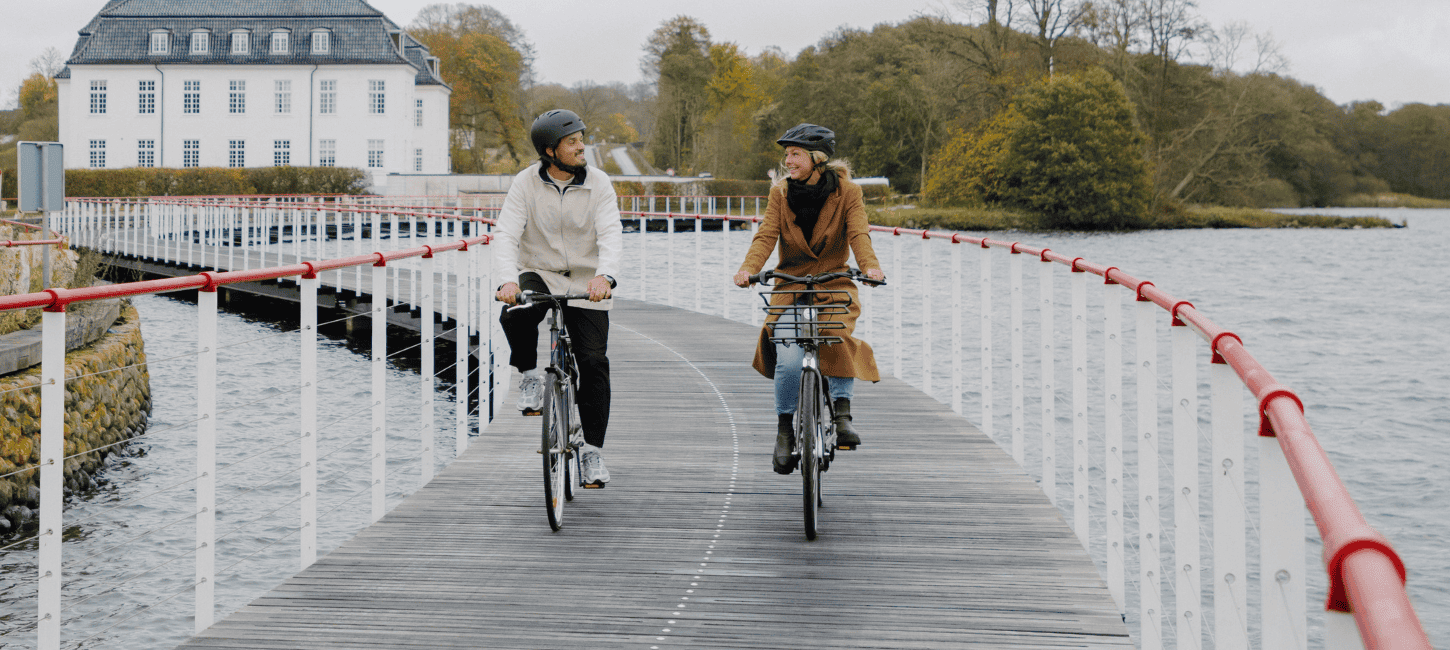 Two cyclists are riding on a bike path built on water, with a white manor house in the background