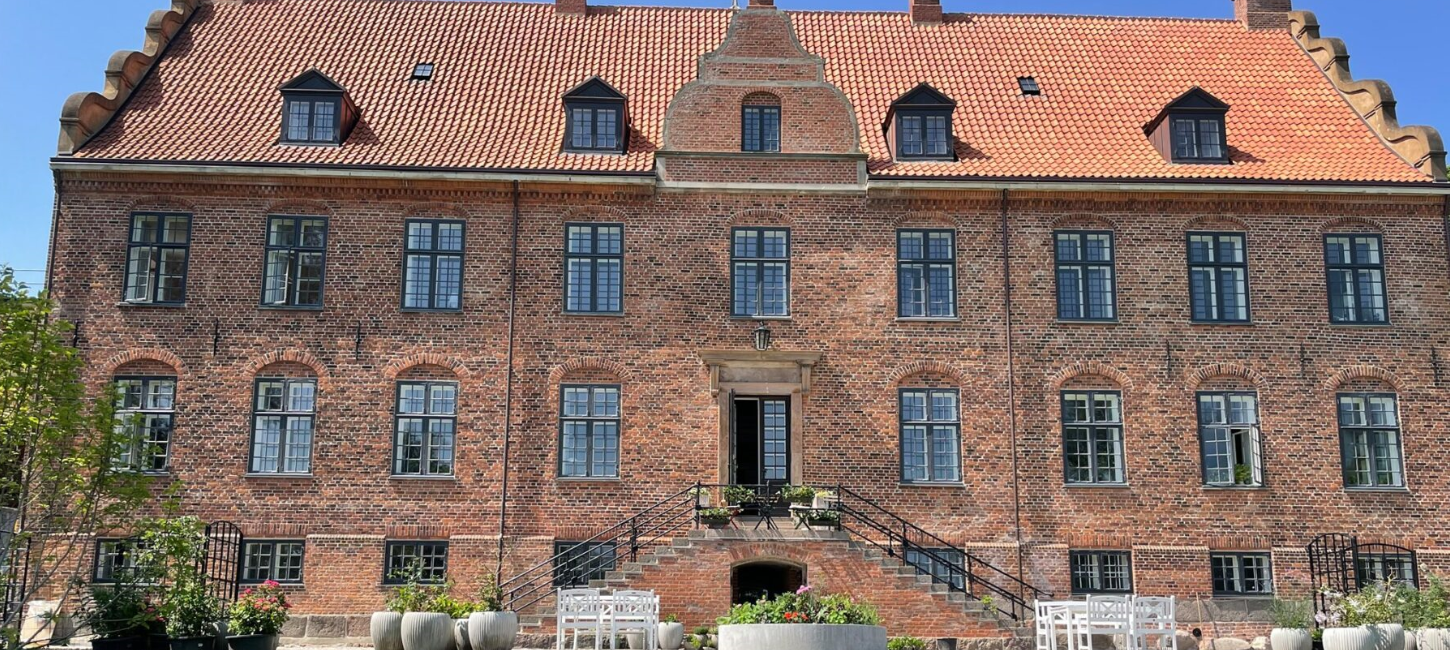 Stor rød herregård med rødt tag og grønne planter i forgrunden.