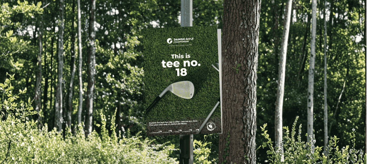 En plakat med teksten "This is tee no 18" hænger i en lygtepæl på en baggrund af træer og grønne blade