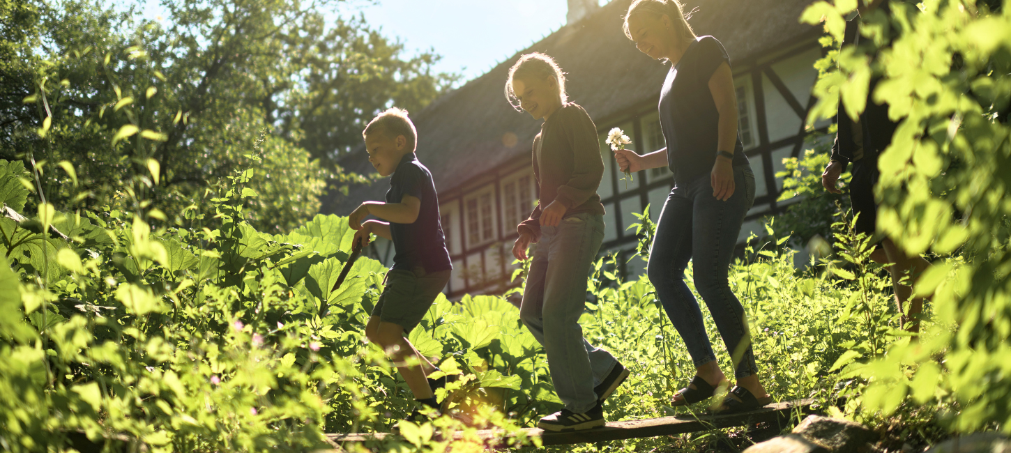 To børn og en kvinde går på et bræt midt i et grønt område med et gammeldags bindingsværkshus i baggrunden. 