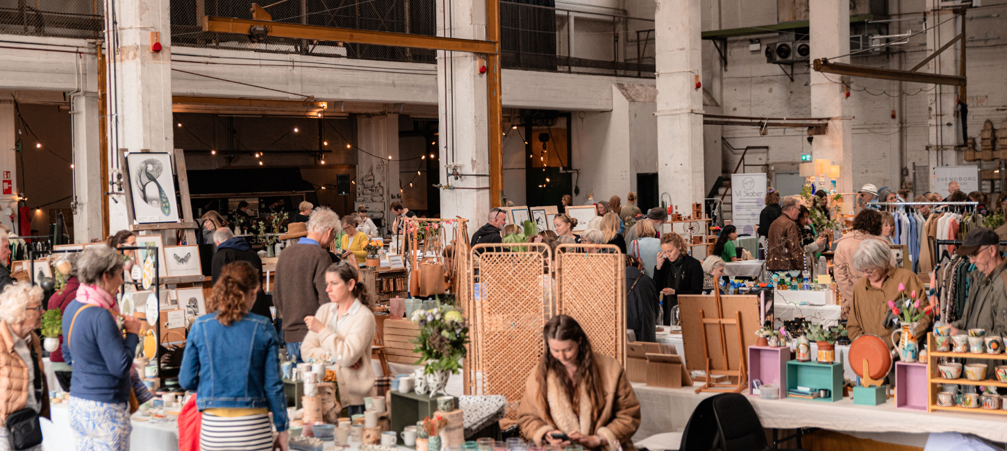 Et marked i en hal med højt til loftet er i fuld gang med mennesker, borde med varer og ting.