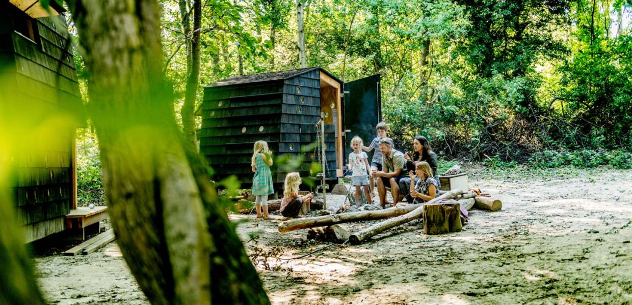 Fem børn og to voksne samlet om et bål i skoven. Bag ved dem ses et arkitekttegnet shelter.