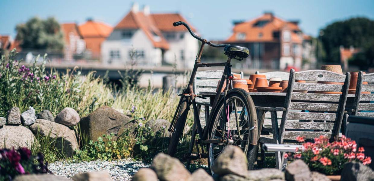 En cykel står lænet op ad et havebord med stole i rustikt træ i vandkanten med hvide huse i baggrunden