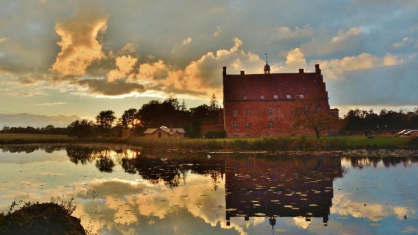 Spejling af det smukke røde slotshotel Broholm Slot i sø med oplyste skyer som baggrund.