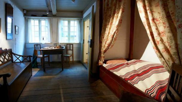 Soveværelse med gammeldags slagbænk af træ, himmelseng med stribet sengebetræk og træspisesæt for to.