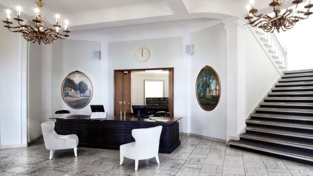 Stilren hotelreception med mork disk, hvide vægge, store hvide lænestole og runde malerier på væggene ved siden af en bred, mørk trappe.