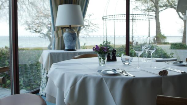 Et rundt bord er dækket med hvid dug, en lille vase med blomster og bestik og vinglas til to. Rundt om bordet står træstole, og gennem det store vindue er der udsigt ud over vandet.