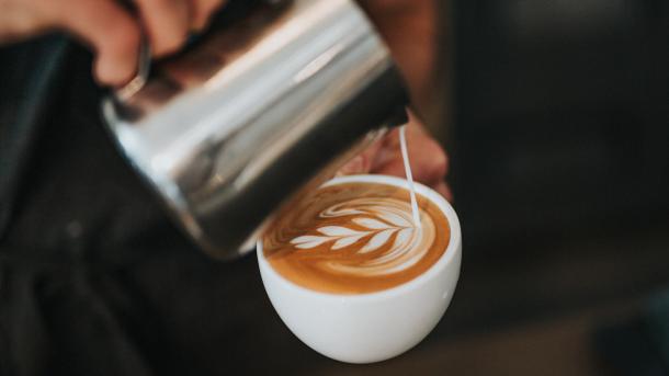 To slørende hænder er ved at lave en kop barista kaffe. I den ene hånd er en hvid kaffe kop fyldt til kanten med kaffe. I den anden hånd er en sølvkande. Fra kanden hældes der mælk ned i koppen som former et blad.