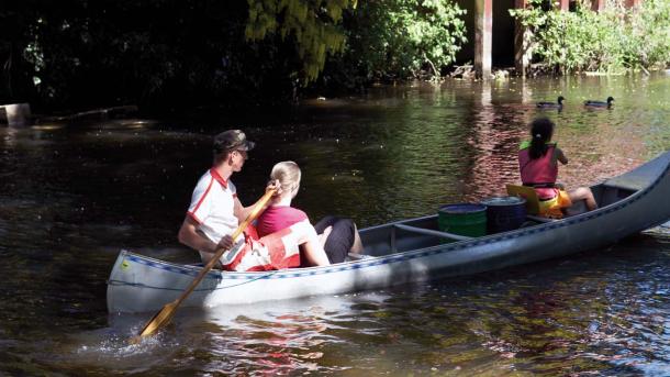 To piger i redningsvest og en mand sejler en grå kano. De sejler på en bred å. I baggrunden ses buske med grønne blade. To ænder svømmer på åen til venstre for kanoen.