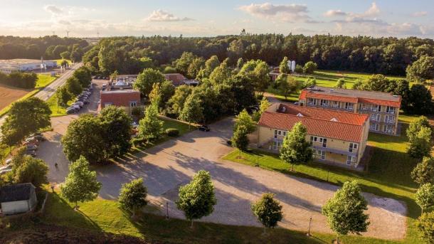 Fjelsted Skov Hotel & Konference set fra luften. En række gul-hvide bygninger med røde tage og masser af fritstående træer omkring grusveje og gårdsplads.