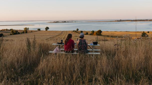 På en mark med udsigt udover havet står et bordebænkesæt. På bænken sidder to kvinder og kigger udover vandet.