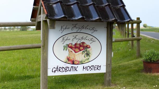 Skilt i vejkanten med et billede af æbler og teksten "Nybro Frugtplantage - Gårdbutik Mosteri"