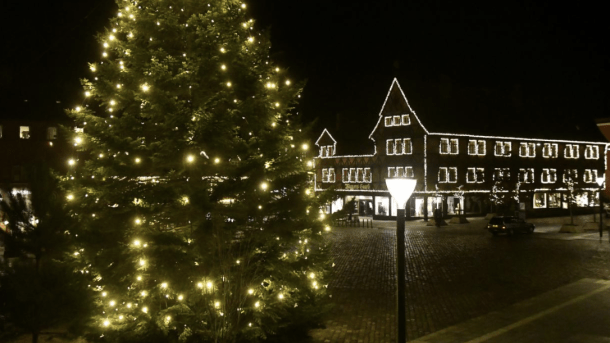Til højre står et sort juletræ lyst op af julelys midt på et torv. Til højre ses en stor bygning hvor vinduerne er markeret med julelys.