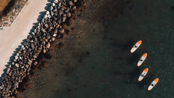 Dronebillede af fire personer på SUP-boards på havet nær en mole dækket af sten