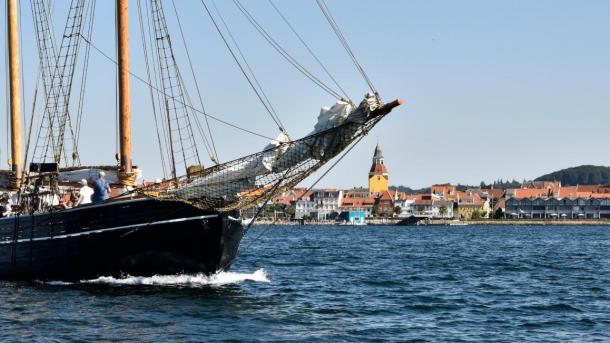 Et tomastet sejlskib i træ sejler på havet foran Faaborg med det karakteristiske kirketårn i baggrunden