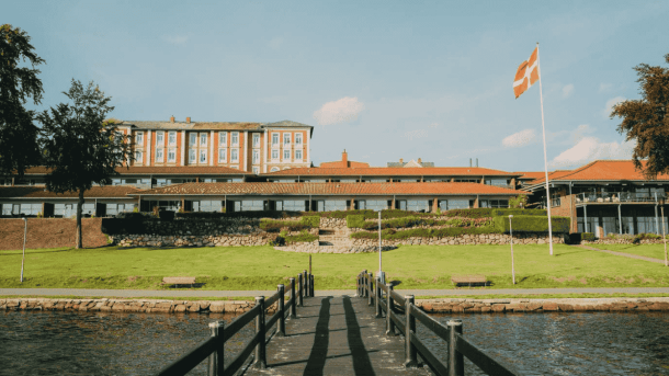 Et rødmuret hotel der ligger på et grønt område lige ned til vandkanten, taget fra en badebro