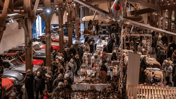 Billedet viser et travlt julemarked i Egeskov på Fyn med et festligt indendørs marked, hvor mange besøgende udforsker de forskellige boder. Markedet er pyntet med julepynt, herunder lysguirlander. Klassiske biler står opstillet langs siderne og tilføjer en unik stemning til den festlige atmosfære. Egeskov danner en livlig ramme om julen på Fyn.