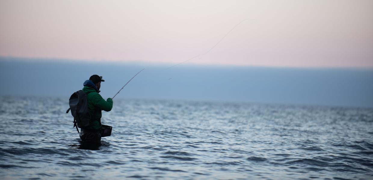 En silhuet af en sportsfisker, der står ude i vandet med ryggen til og fisker. Han har kasket på og rygsæk. Grå og grumset himmel og horisont.