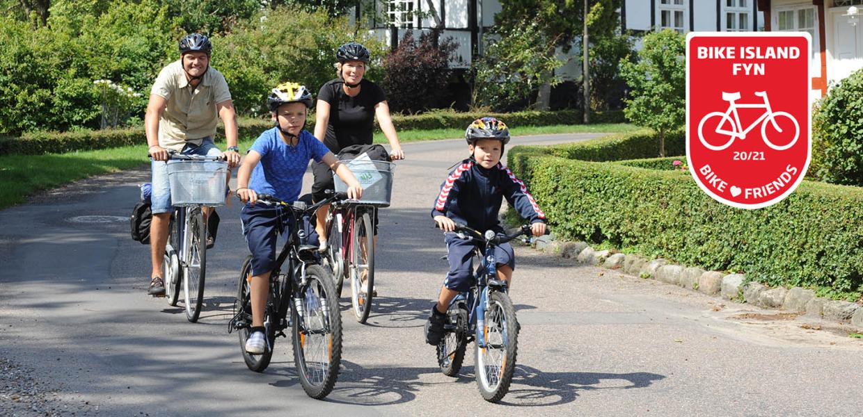 To voksne og to børn cykler af sted på stille asfaltvej med bindingsværkshus i baggrunden. Øverst i højre hjørne ses det røde Bike Friend-logo.