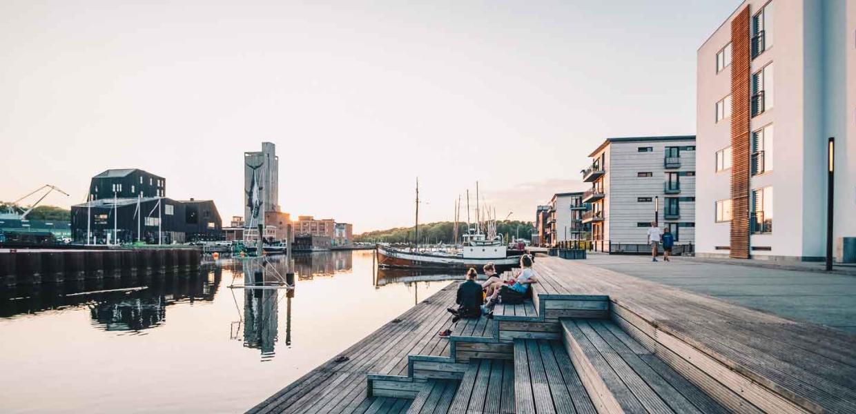 Stemningsbillede fra Odense havn en aften med solnedgang. Mennesker sidder på en træplatform ud til vandet og kigger på skibene.