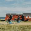 To cyklister kører på en grusvej med græs til den ene side og hav og badehuse til den anden side