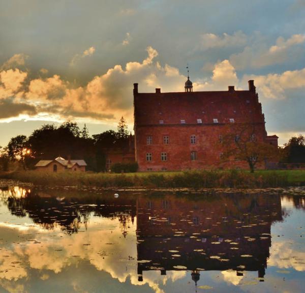 Spejling af det smukke røde slotshotel Broholm Slot i sø med oplyste skyer som baggrund.