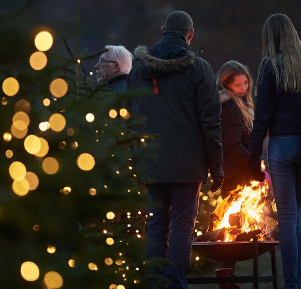 Et par mennesker omkring ild i et bålfad i mørket. Lyskæde på træ i forgrunden.