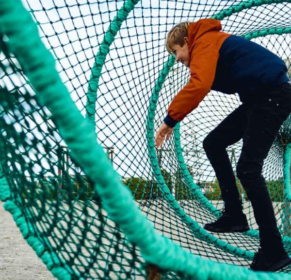 Et større barn inde i 'fiskeruse' på en legeplads med faldsand. I baggrunden ses et legetårn.