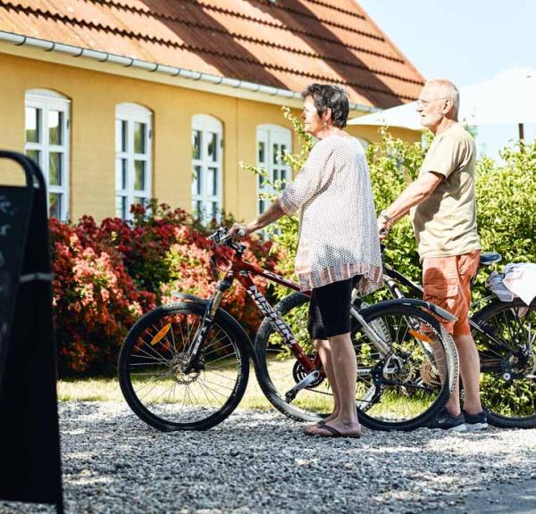 Mand og kvinde står afventende med deres cykler ved gult hus. 