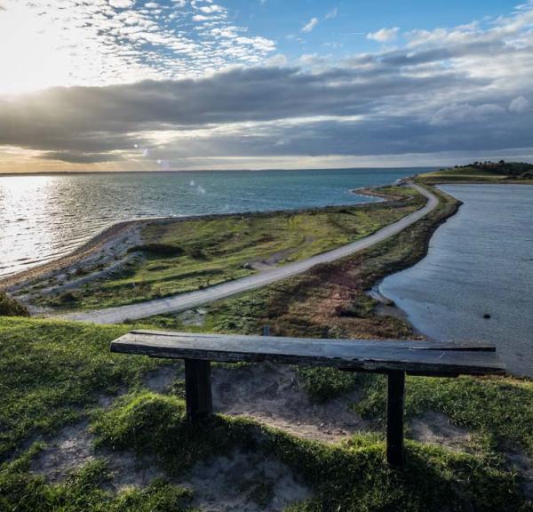 Udsigt over tange med smal vej og vand på begge sider. Solen stråler dramatisk gennem lavthængende grå skyer. I forgrunden står en træbænk.