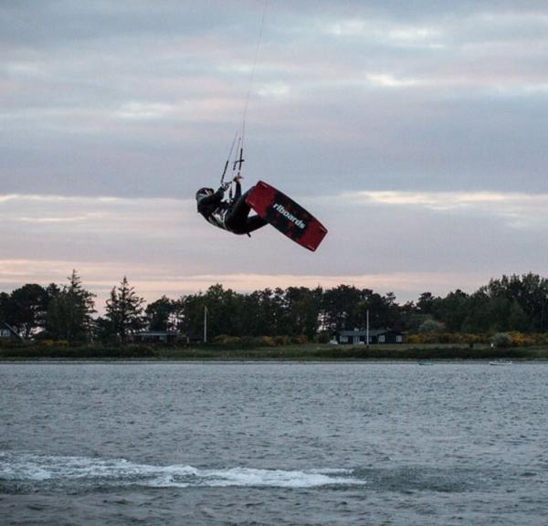 Midt i billedet flyver en kitesurfer over havet. Til venstre i billedet kan man se en sejlbåd på vej ud af billedet. Til højre er endnu en kitesurfer. Han surfer på vandet. I horisonten kan man se en skov på land. 