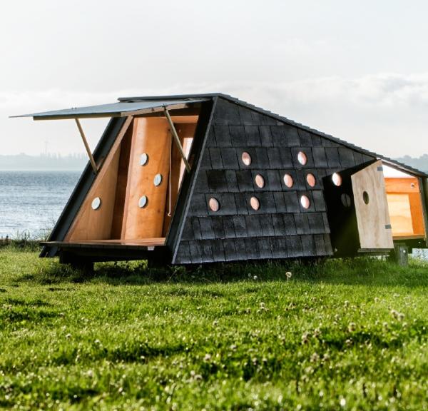 Shelter med åbne døre, der afslører et varmt interiør i træ. Det befinder sig i grønne omgivelser med udsigt over havet.