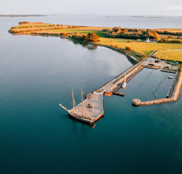 Dronefoto af Skarø havn, hvor en tomastet skonnert ligger som den eneste båd for enden af havnen