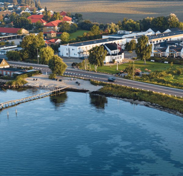 Dronefoto af hotel lige ud til kysten med grønt græs, blåt hav og et rødt svajende dannebrog