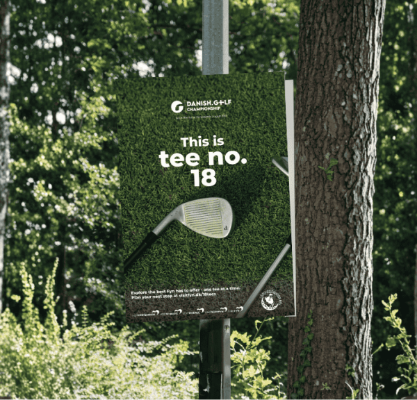 En plakat med teksten "This is tee no 18" hænger i en lygtepæl på en baggrund af træer og grønne blade