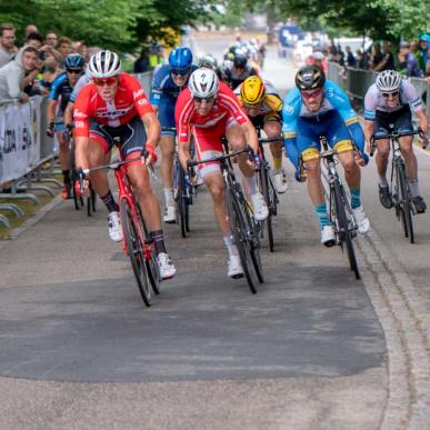 Cykelryttere på vej ind ad opløbsstrækningen i tæt løb. Tilskuere står bag hegn og hepper til begge sider.