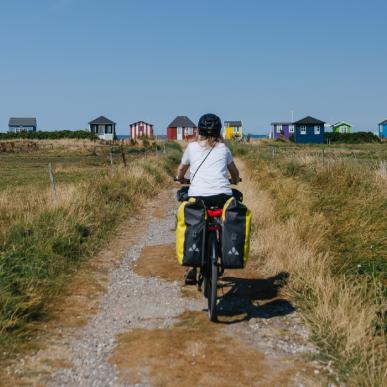 Kvindelig cyklist ses bagfra på vej ud til badehusene ved Vester Strand på Ærø. Det er sommer.