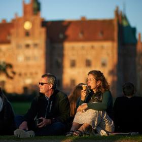 Et par mennesker sidder på græsset udenfor i aftensolen. I baggrunden ses Egeskov Slot.