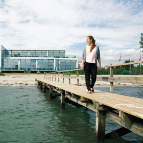 En dame går langs en badebro og kigger ud over vandet. Et stort hotel med vinduer på hele facaden ligger i baggrunden.