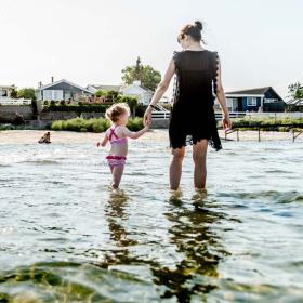 En mor går med en lille pige i hånden og sopper i vandet langs strandkanten.