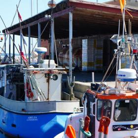 En rød murstensbygning står på en havnekaj. Foran ligger et par fiskebåde tøjret fast til kajen. En mand går rundt og kigger på bygningen.